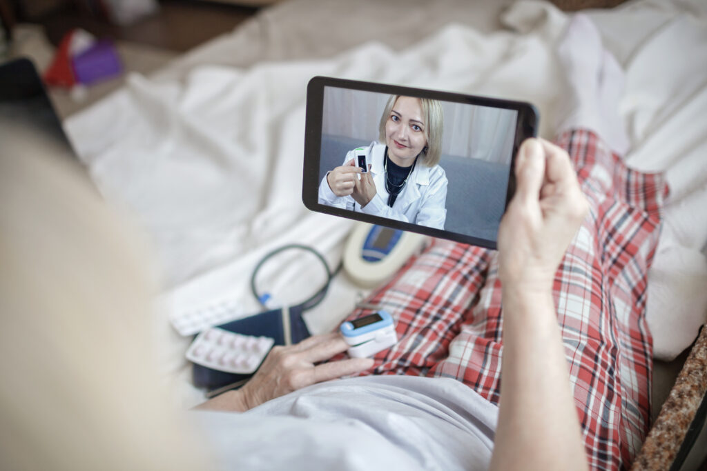 A person lying in bed views a doctor on a tablet screen, who is holding up a medication bottle, with RPM medical supplies in the foreground.